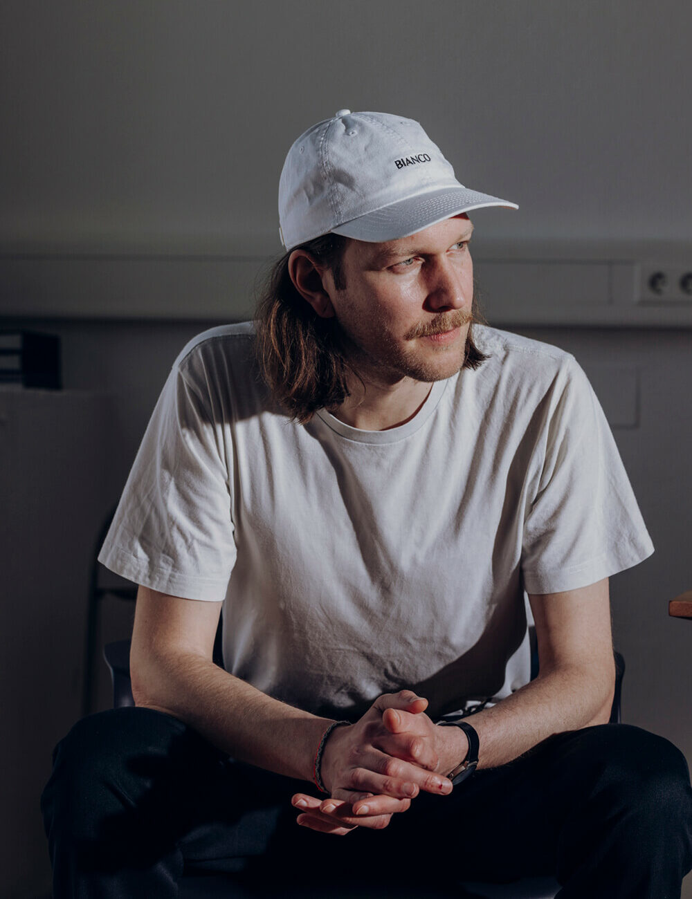 Portrait von Zeno, der mit gefalteten Händen auf einen Stuhl sitzt und ernst an der Kamera vorbeischat. Beiges T-Shirt und weiße Cap.