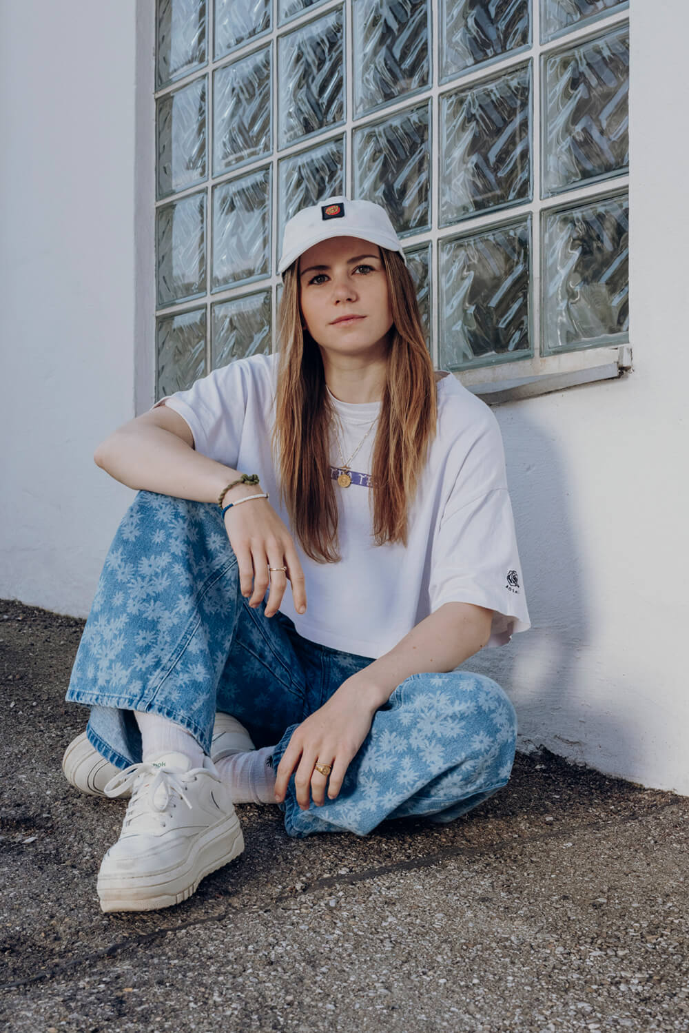 Portrait Nina, die mit verschränkten Beinen locker an einer Hauswand sitzt. Jeans mit blumen, weißes Shirt mit weißer Cap.
