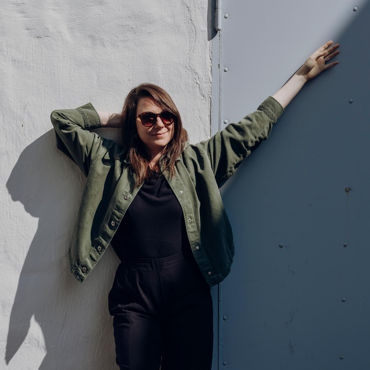 Portrait von Anke die in einem grünen Hemd fröhlich an eine Wand anlehnt und eine Sonnenbrille trägt.