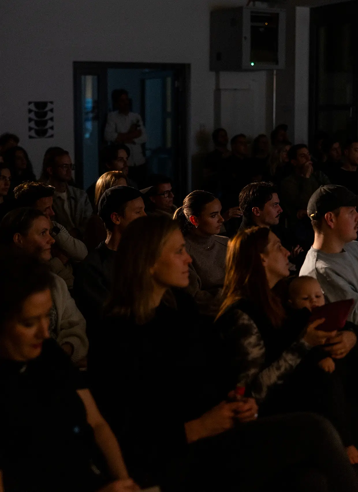 Blick in Zuschauermenge, vorne sitzende, hinten stehende Personen in einem dunklen Raum, Lichtschein von hinten